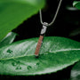 "Campania Necklace" necklace featuring a walnut and Damascus pendant on a chain, set against a lush green leaf background.