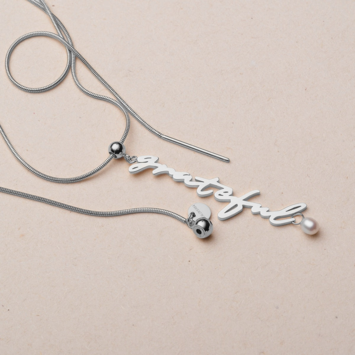 "Grating- Charm" halskæde med elegante perler og sølv, byder på en delikat charme og stilfuld script design.